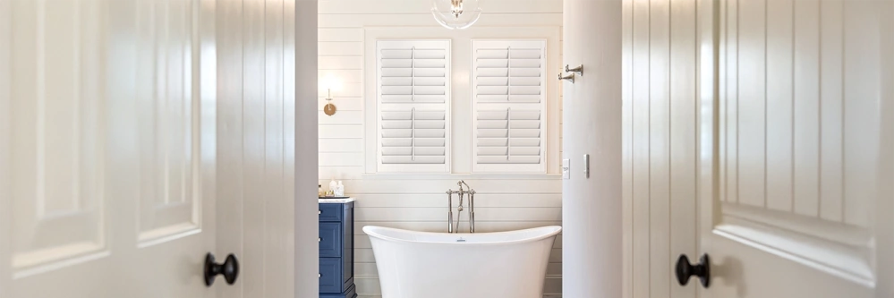 White Polywood shutters on two windows over a freestanding bathtub